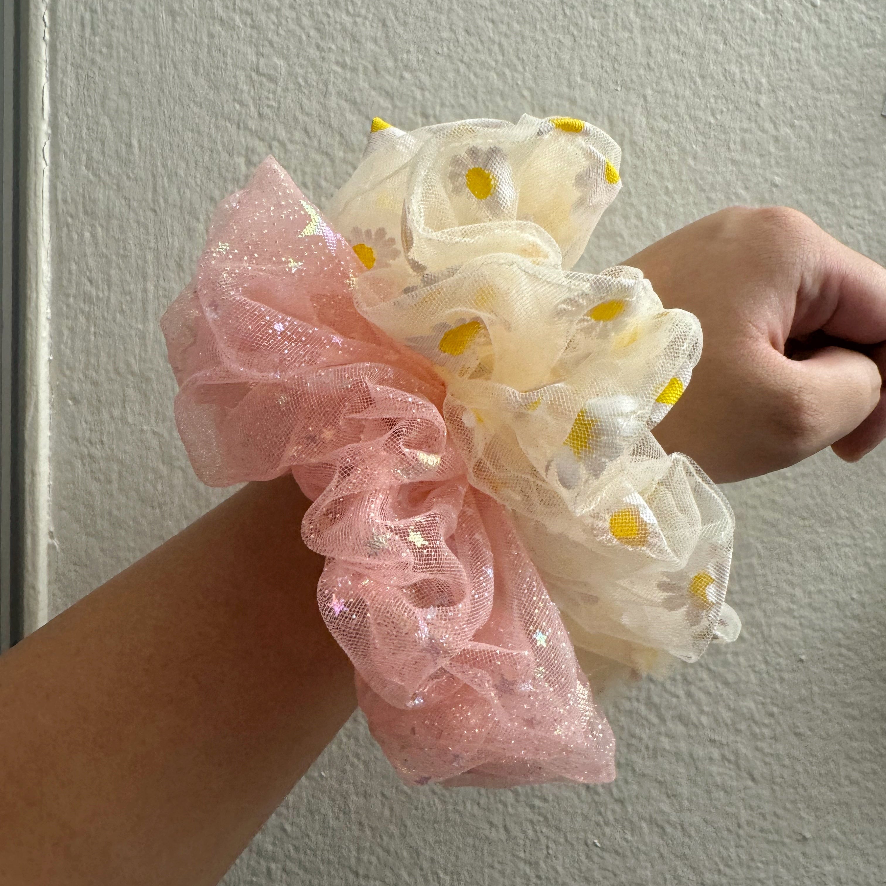 Two scrunchies, one pink and one yellow with yellow daisy patterns, held against a plain background.