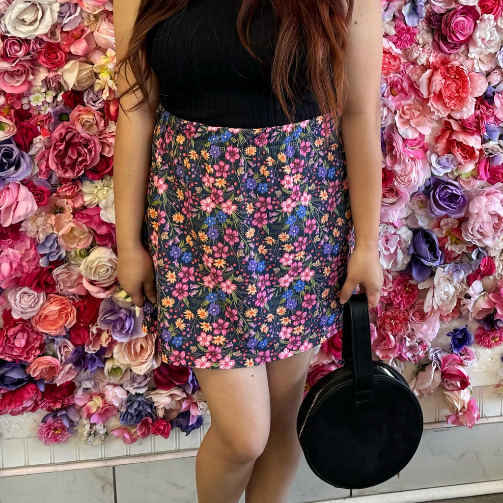 Model wearing a black top with our Mila floral skirt standing in front of a colorful flower wall.